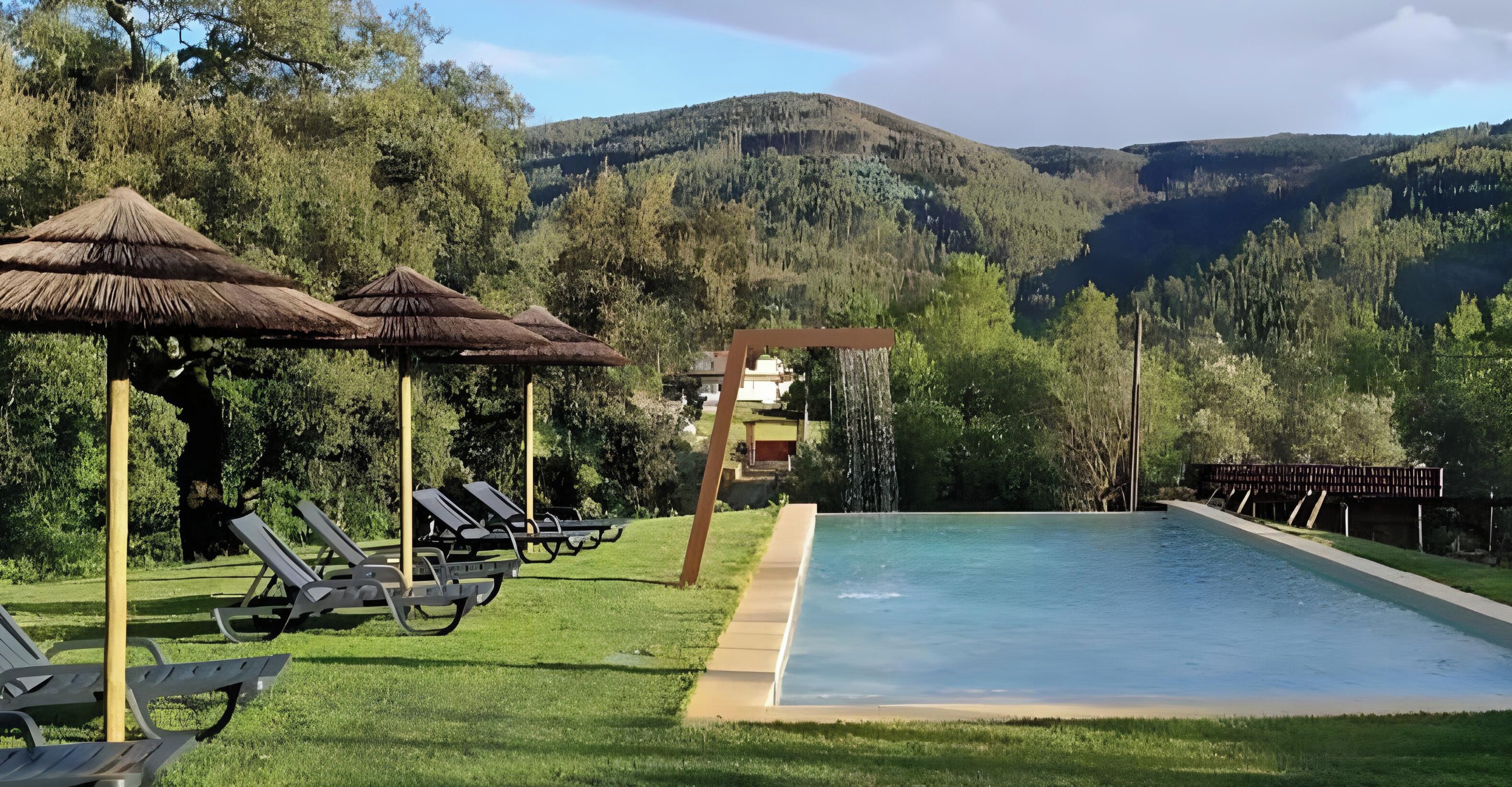 Une piscine entourée de chaises longues et de parasols, elle-même bordée d’une pelouse et d’arbres, avec une montagne en toile de fond.