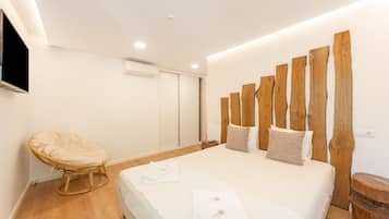 Une chambre à coucher comprenant un lit, une tête de lit en bois, un fauteuil en osier et un téléviseur à écran plat fixé au mur.