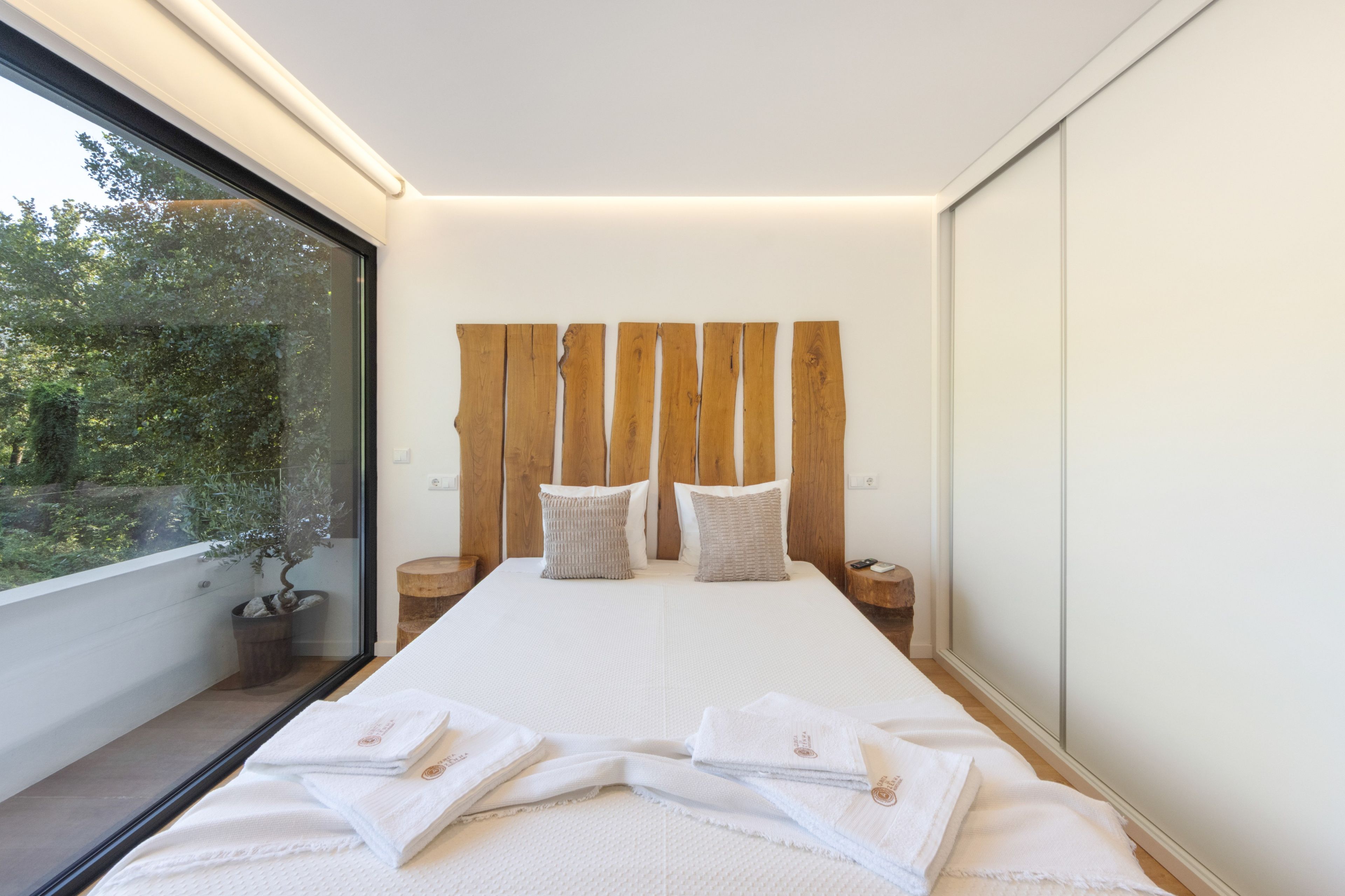 Une chambre moderne avec un grand lit, une tête de lit en bois, deux tables de chevet et une vue sur la verdure par la fenêtre.