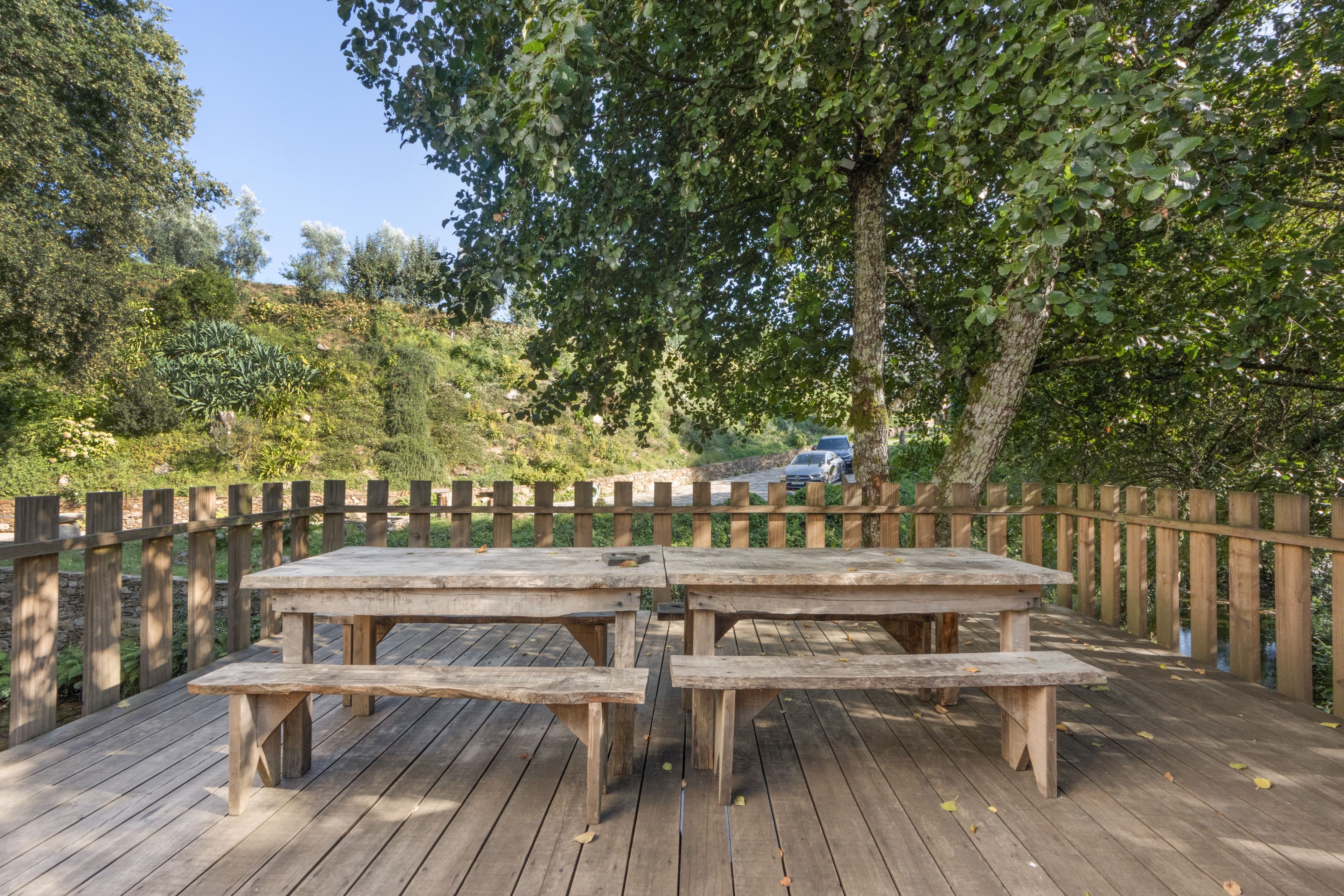 A wooden deck with picnic tables and benches, surrounded by a wooden fence, with trees and a car in the background.