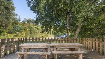 A wooden deck with picnic tables and benches, surrounded by a wooden fence, with trees and a car in the background.
