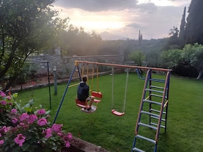 A child on a swing set in a grassy backyard with a stone wall and trees in the background.