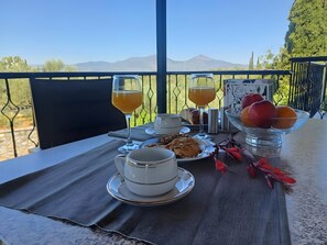 Un tavolo apparecchiato con una tazza, un bicchiere di succo, un piatto di biscotti, una ciotola di frutta e un cornetto su una terrazza con vista sulle montagne.