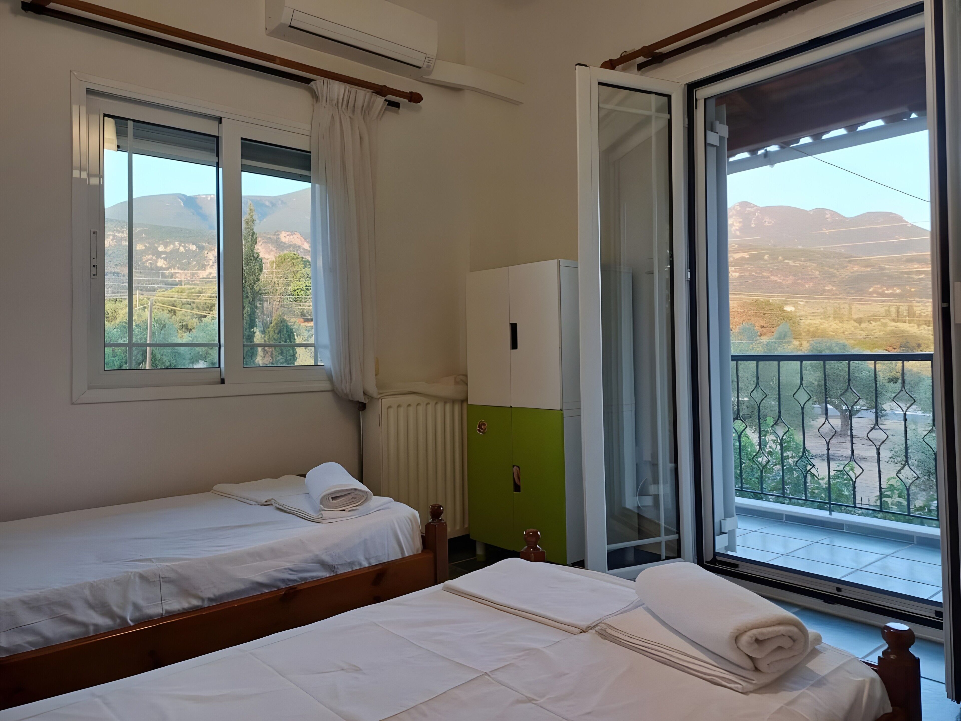A bedroom with two beds, a radiator, a window with curtains, and a balcony with a view of the countryside.