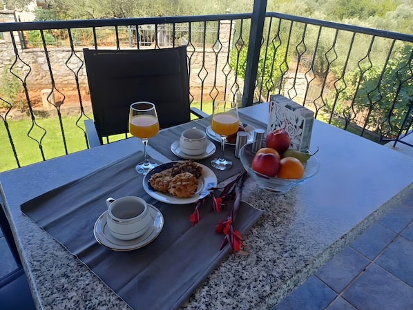 A table with breakfast items, a glass of orange juice, and a bowl of fruit on a balcony with a view of greenery.