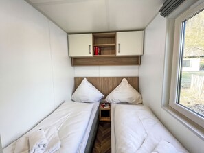 Une chambre de taille modeste, équipée de deux lits simples, d’une petite étagère en bois où sont posés des livres et d’une fenêtre donnant sur l’extérieur.