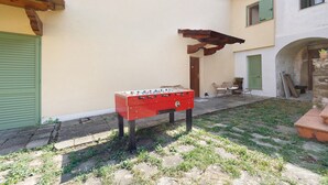 A red foosball table in a courtyard with a stone floor, a wooden pergola, and a building with green shutters.