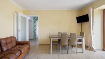 Ein Wohnzimmer mit braunem Ledersofa, einem Esstisch mit Stühlen, einem an der Wand befestigten Flachbildfernseher und einem Flur mit einer Tür zu einem weiteren Raum.