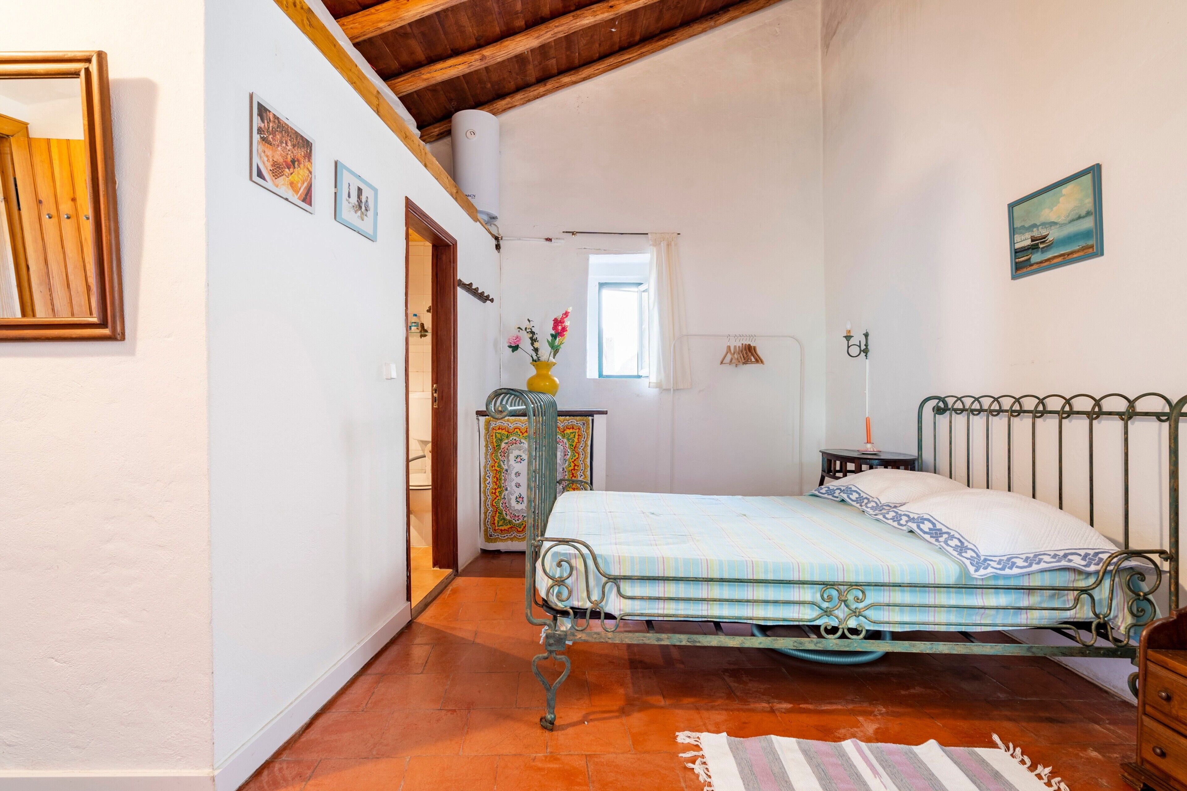 A bedroom with a metal bed frame, a wooden dresser, a small table with a lamp, and a window with a view of the outside.