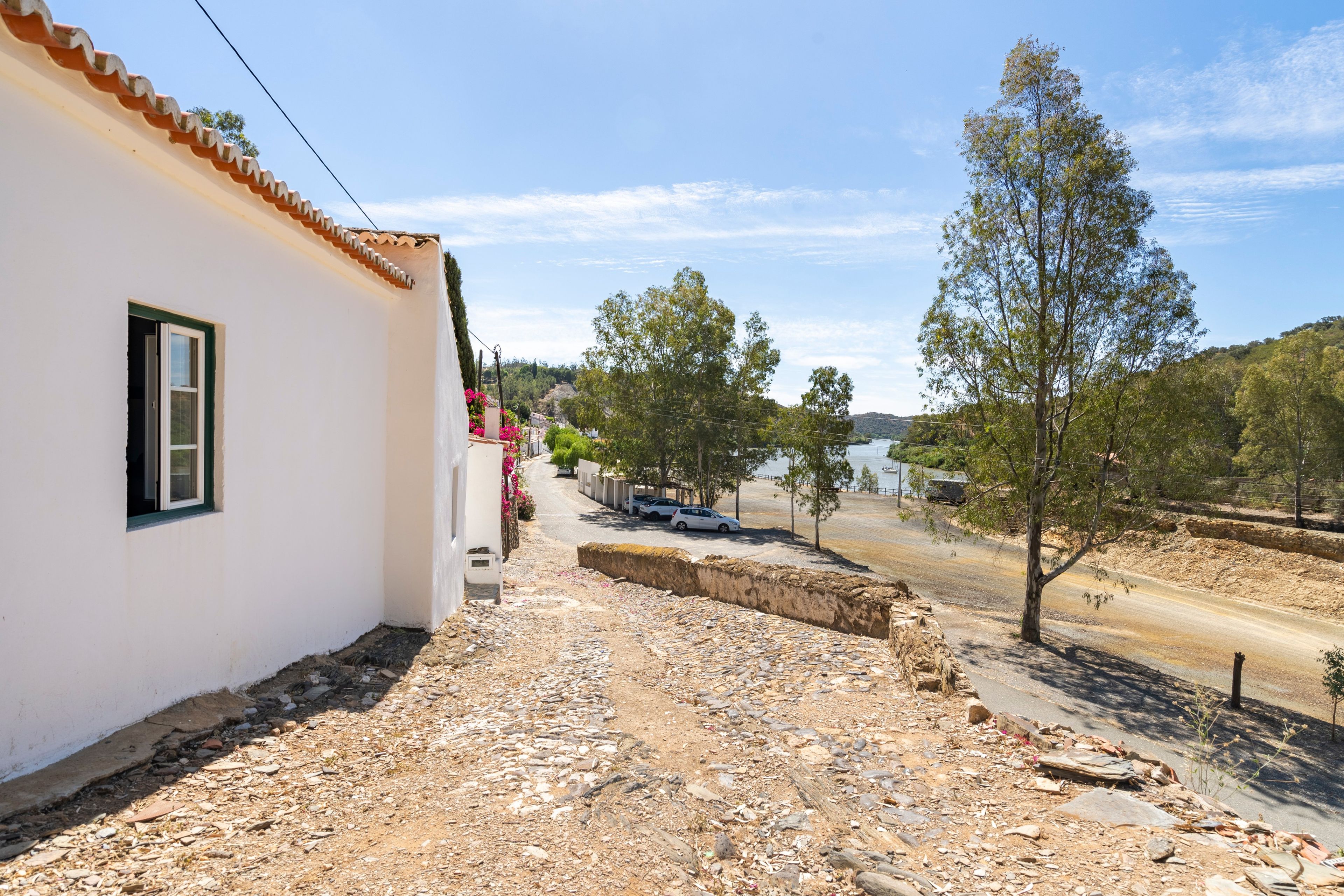 Une rue étroite, avec un bâtiment blanc sur la gauche, un chemin de terre et des arbres sur la droite.