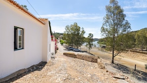 A narrow street with a white building on the left, a dirt path, and trees on the right.
