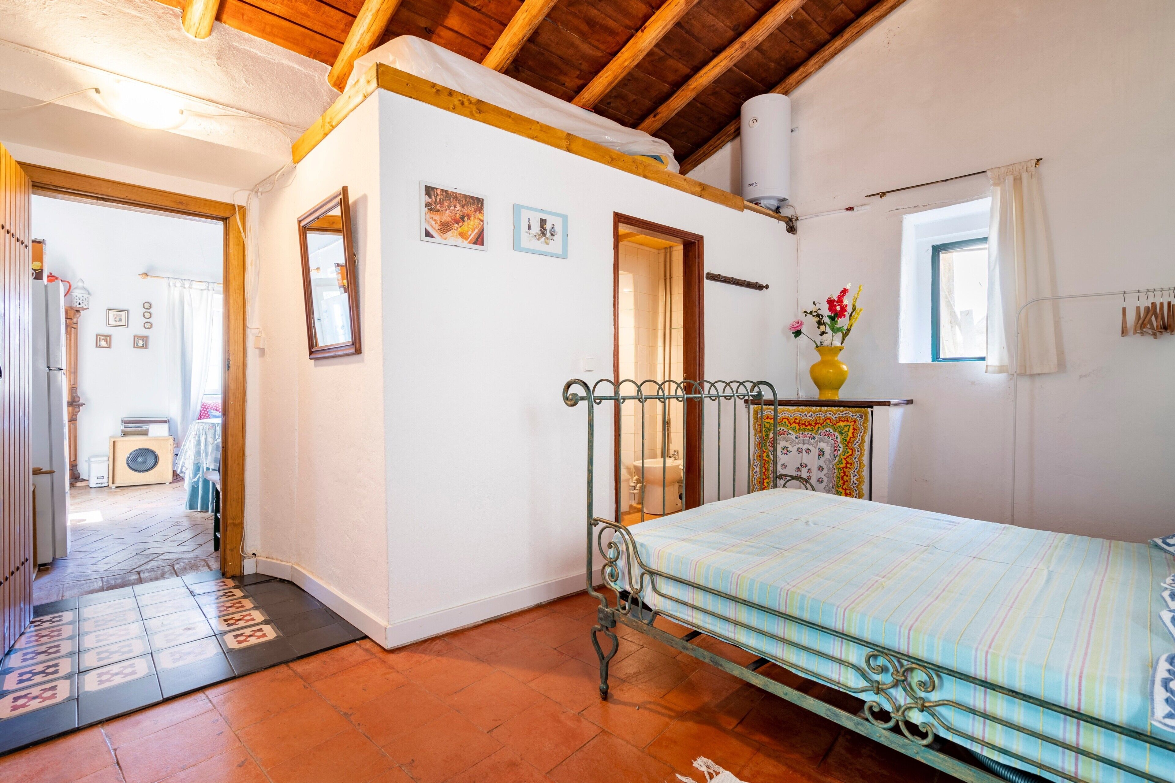 Un dormitorio con estructura de cama de metal, una mesita de noche con un florero y una ventana con cortinas.