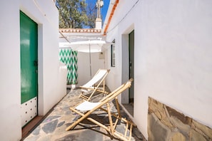 A narrow corridor with white walls, a green door, and a tiled floor. There are two wooden lounge chairs and a white umbrella.