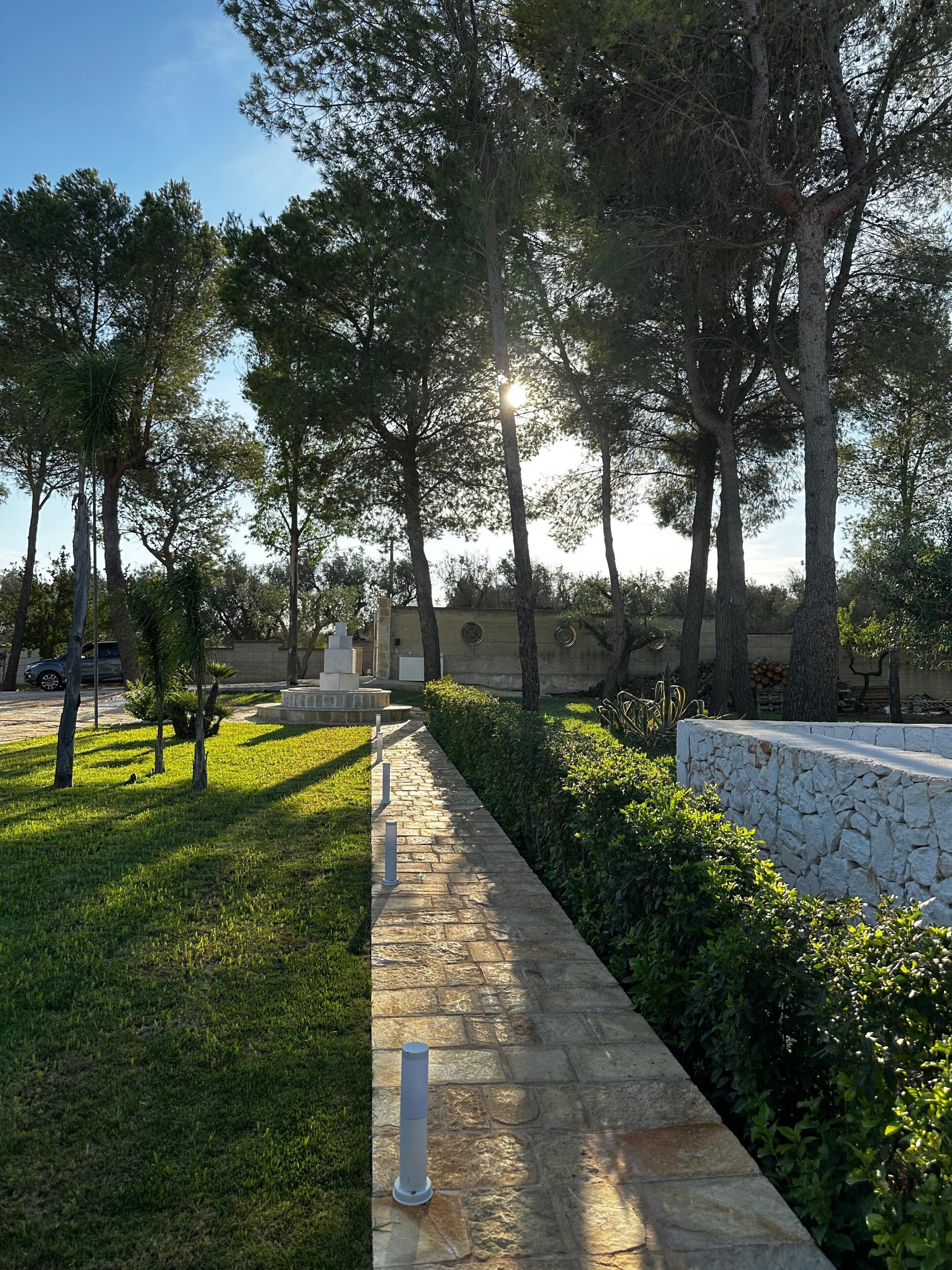 Un chemin bordé d’arbres et flanqué d’un mur de pierres, qui mène à un bâtiment à la façade en pierre.