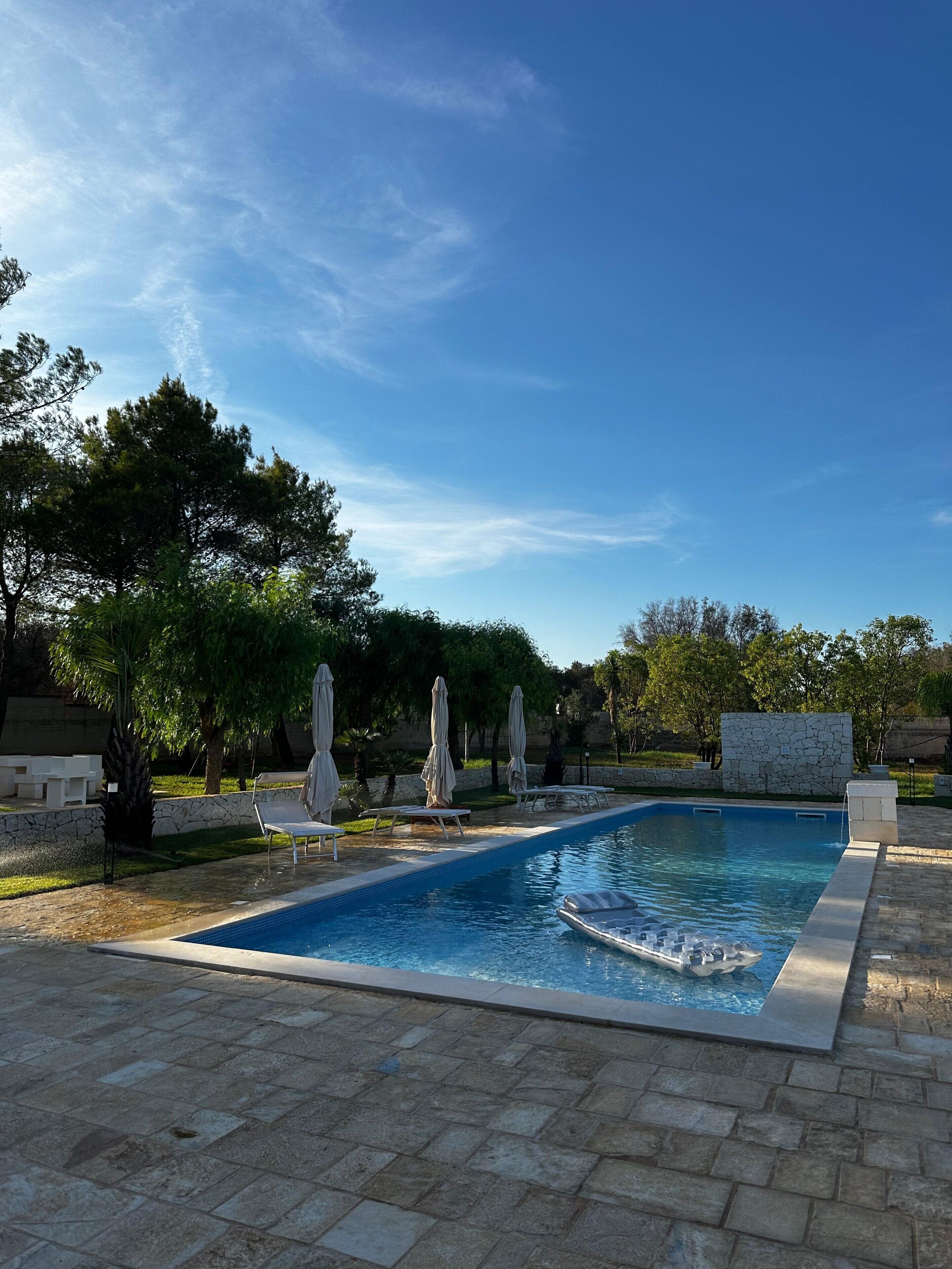 Une piscine entourée d’arbres et d’un mur de pierre, avec des chaises longues et des parasols à proximité.