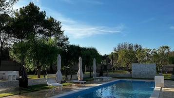 Une piscine entourée d’arbres et d’un mur de pierre, avec des chaises longues et des parasols à proximité.
