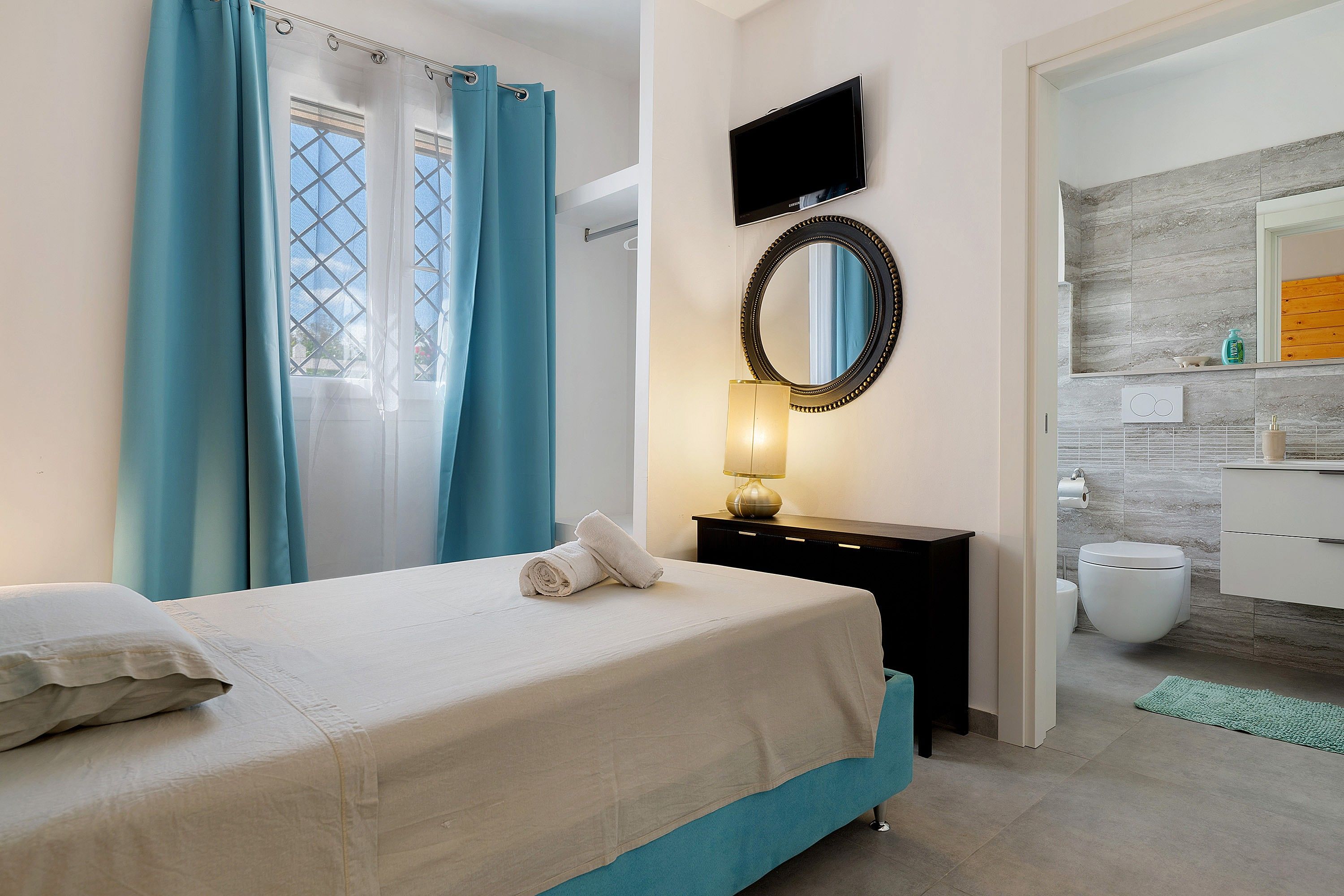 Un dormitorio con cama, una mesita de noche con lámpara, televisor montado en la pared y un baño visible a través de una puerta abierta.