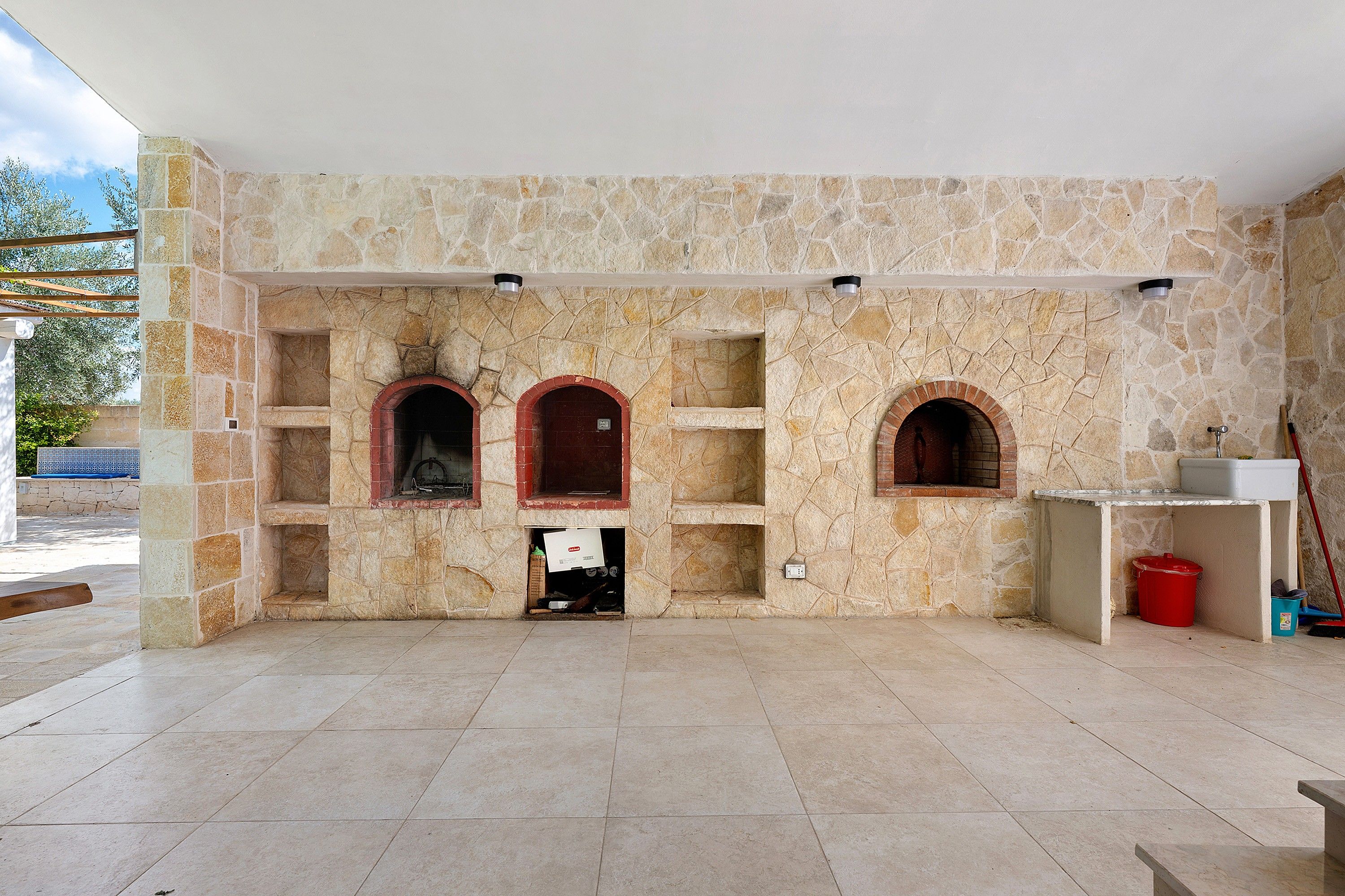 A stone fireplace with three arched openings, a tiled floor, and a red bucket on the right.