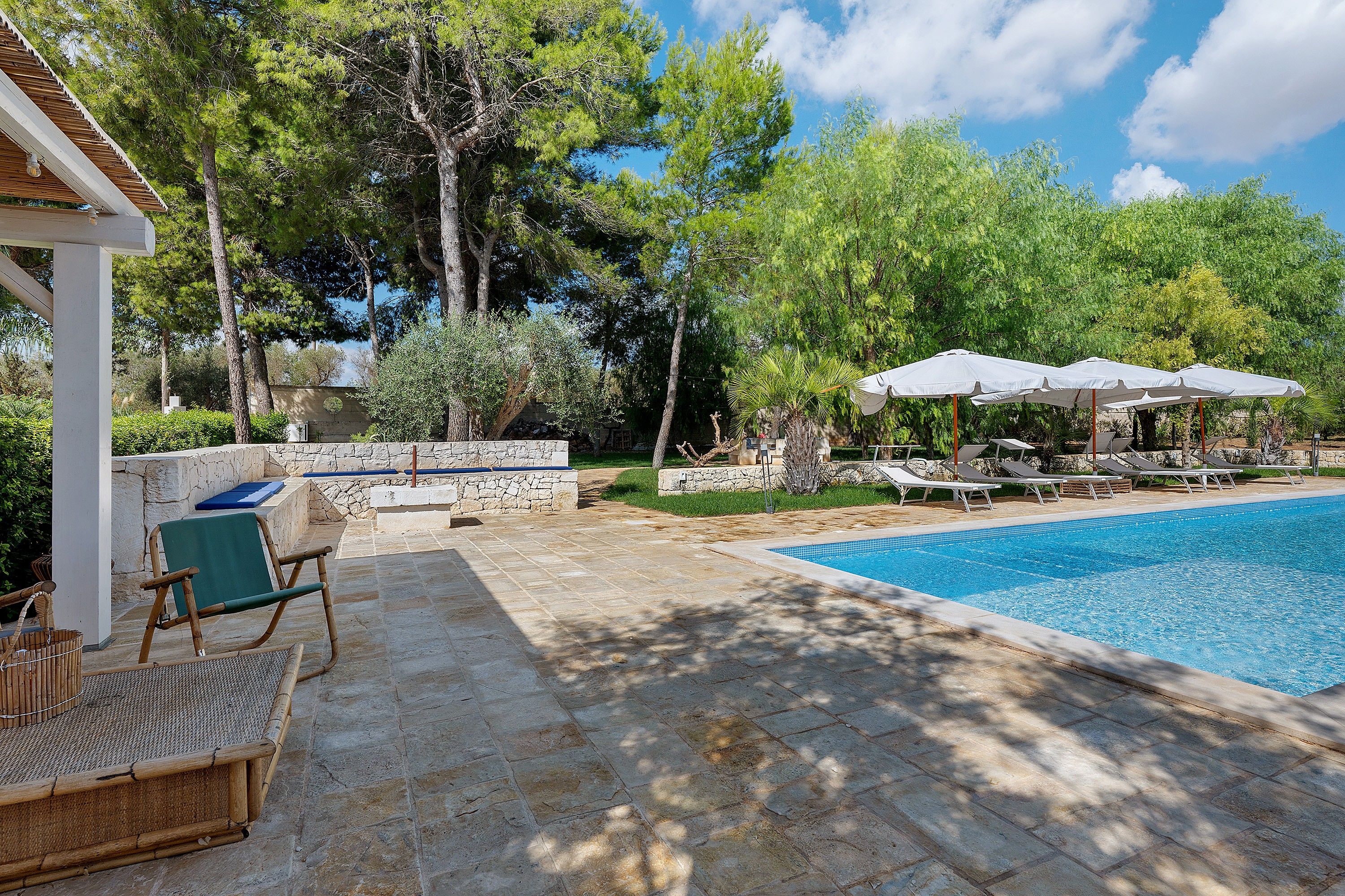 Une piscine entourée d’arbres et d’un mur de pierre, avec des chaises longues et des parasols à proximité.