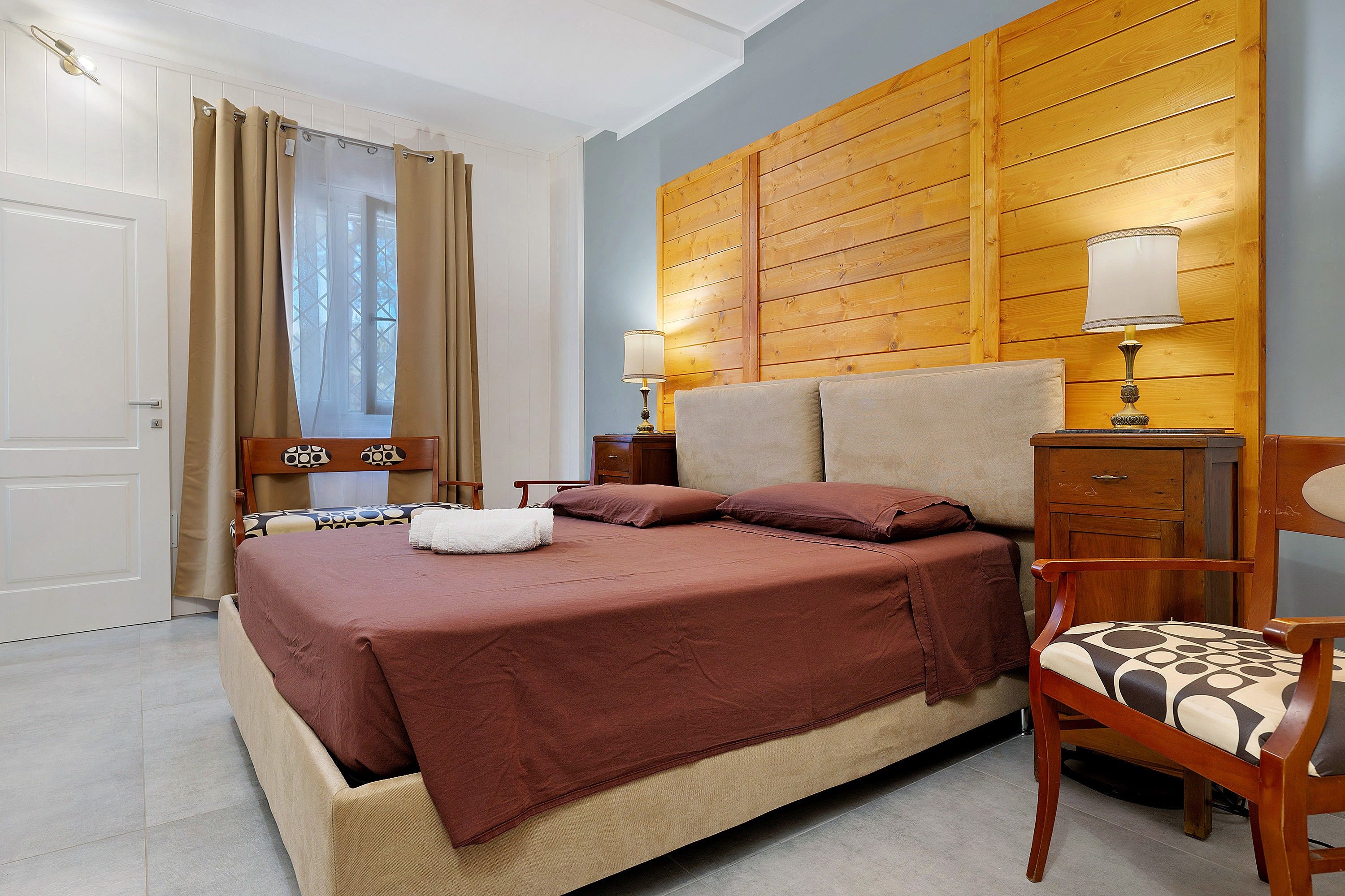Une chambre à coucher avec un lit, une tête de lit en bois, une table de chevet avec une lampe, une chaise et une fenêtre avec des rideaux.