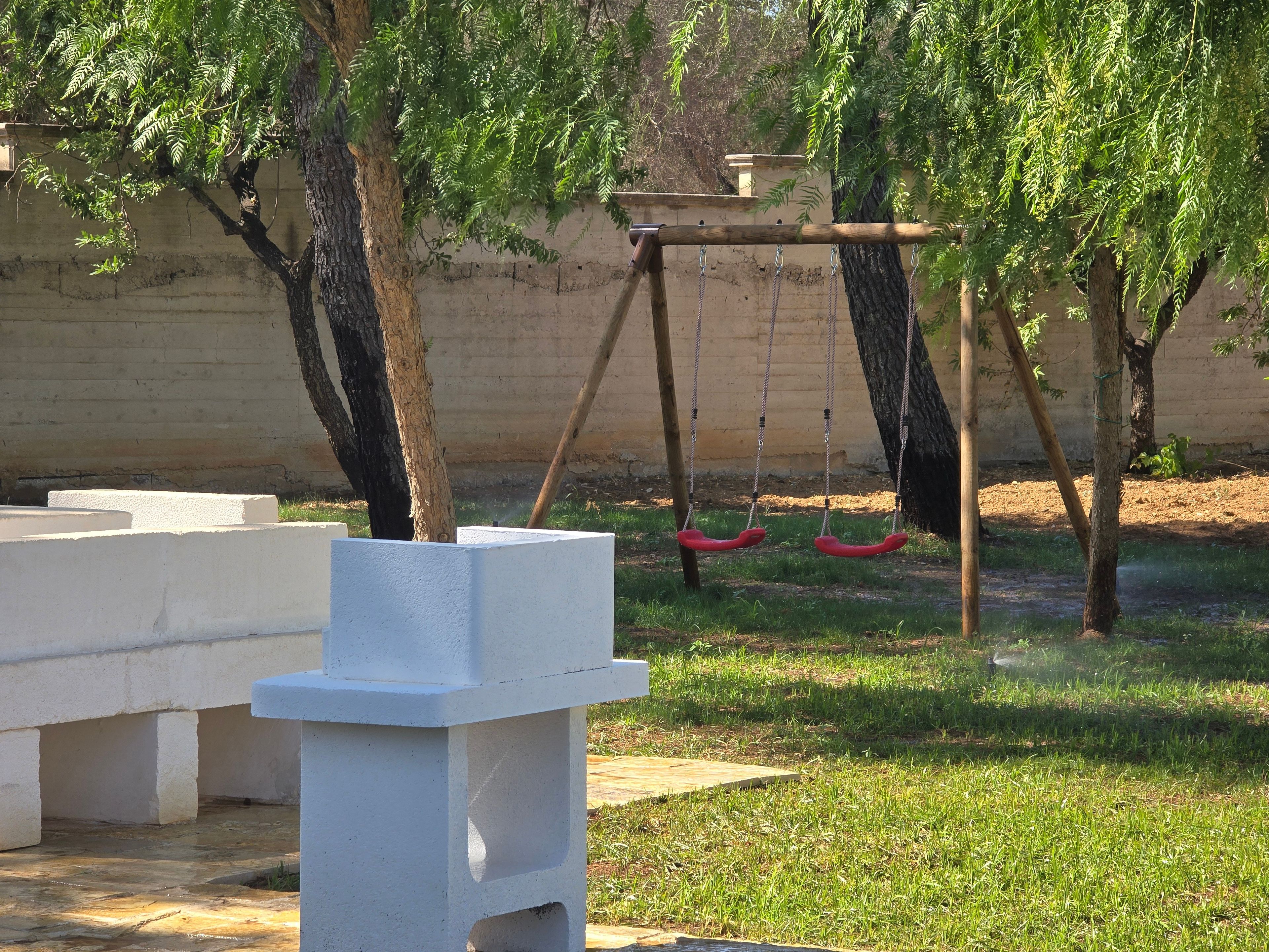 Zona de jocs amb cadenes, una estructura blanca, gespa i arbres al voltant.