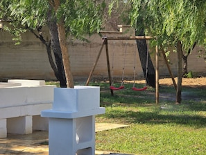 A playground with swings and a white structure, surrounded by grass and trees.
