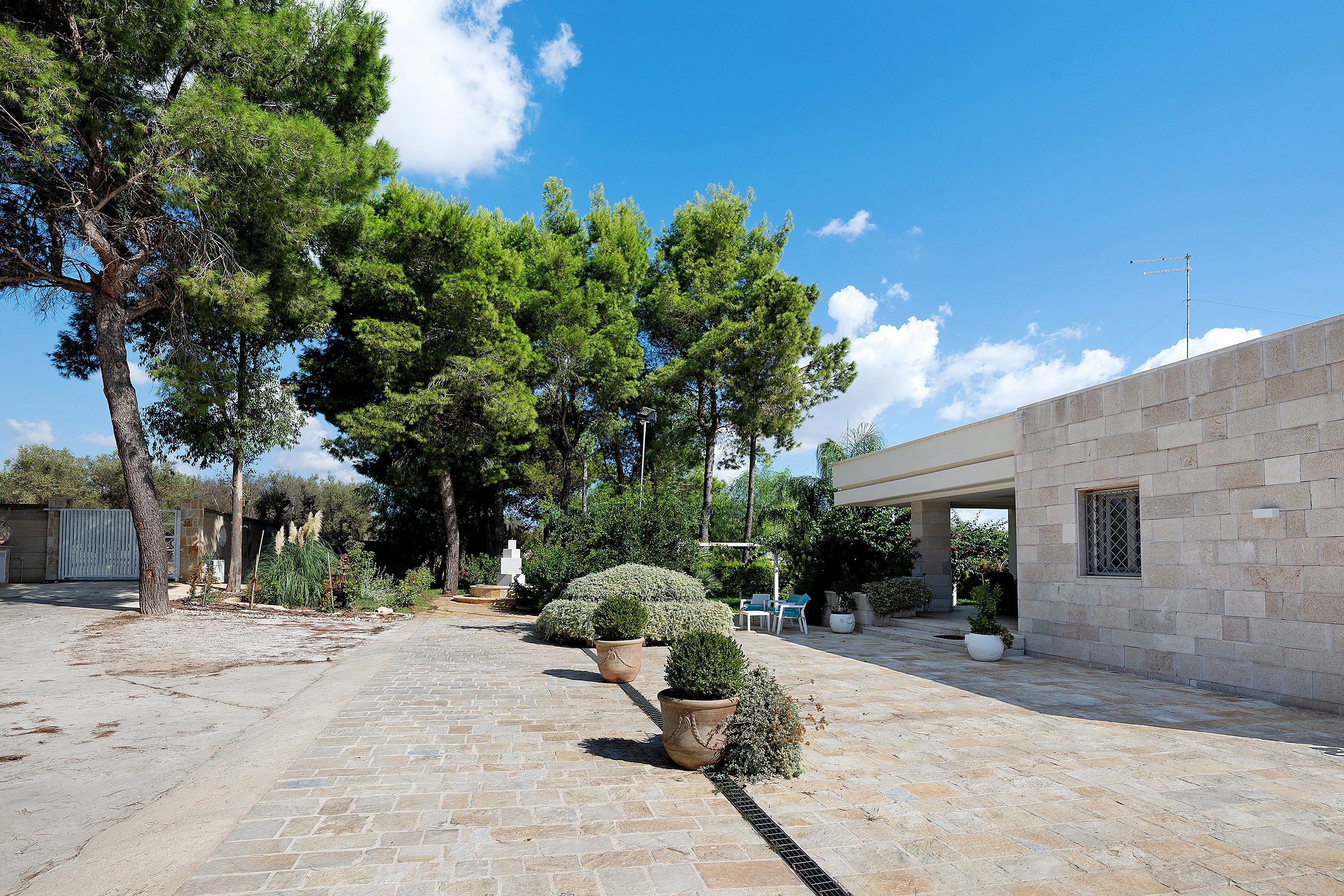 Un cortile pavimentato con piastrelle in pietra, circondato da alberi e piante in vaso, che conduce a un edificio con facciata in pietra.