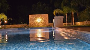 Une piscine nocturne avec un sol carrelé, un mur en pierre agrémenté d’un luminaire et des palmiers en arrière-plan.