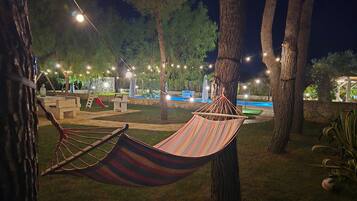 Un'amaca sospesa tra gli alberi in una zona esterna ben illuminata, con piscina e lettini.
