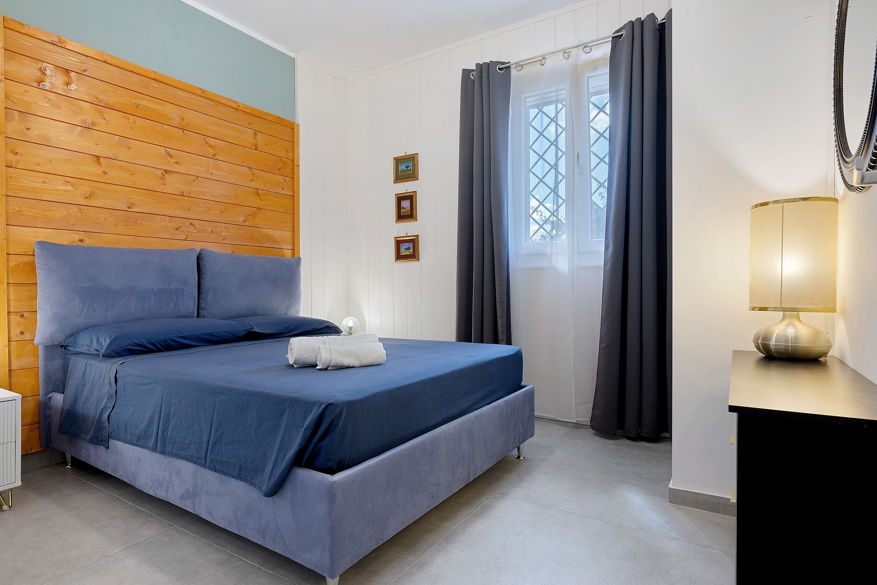 Une chambre à coucher avec une tête de lit en bois, un lit avec une literie bleue, une table de chevet avec une lampe et une fenêtre avec des rideaux.
