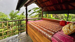 A wooden deck with a bed featuring a patterned bedspread, surrounded by lush greenery and a wooden railing.