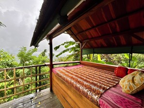 Um deck de madeira com uma cama coberta por um cobre estampado, cercado por vegetação exuberante e um corrimão de madeira.