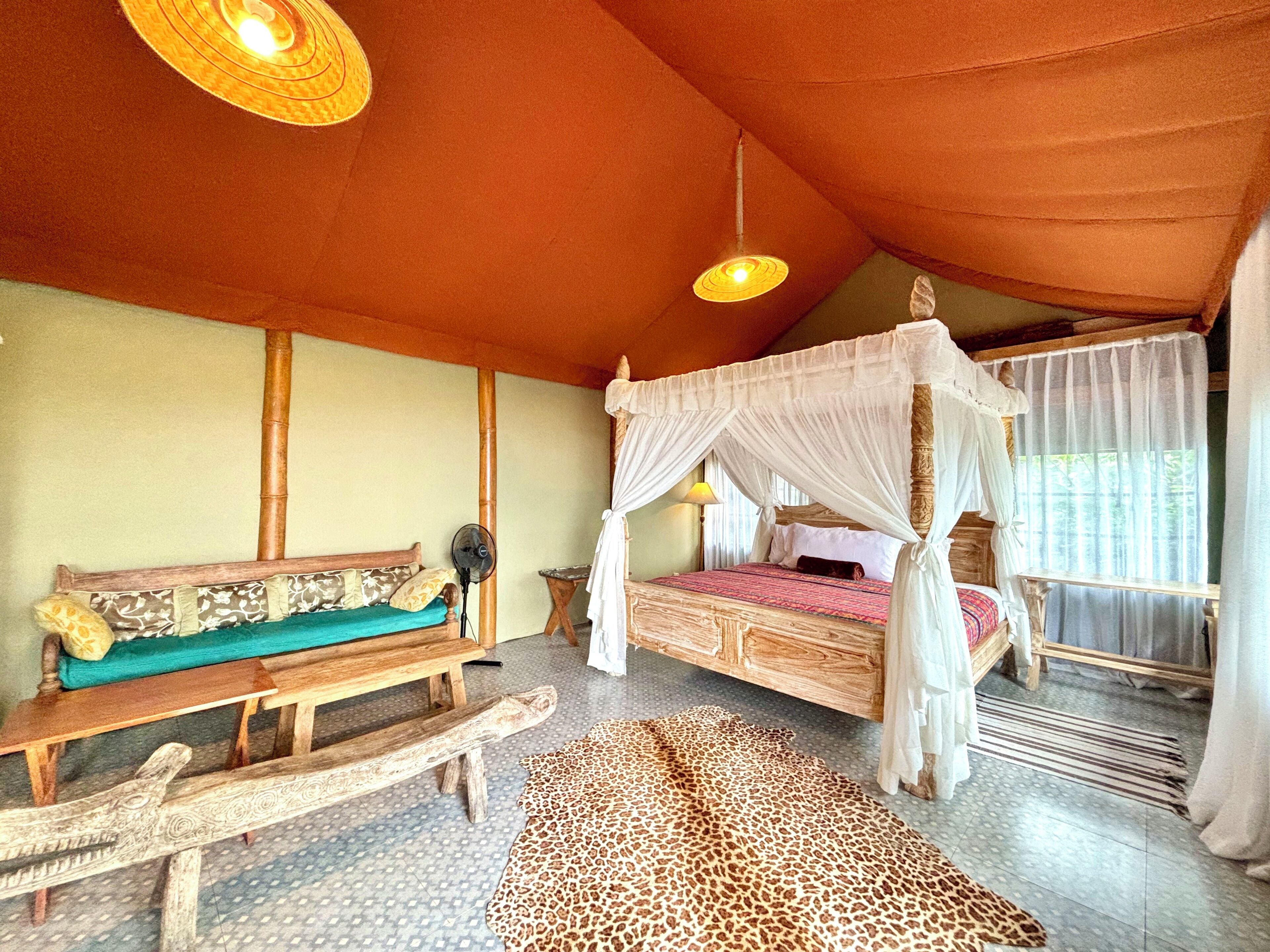 Une pièce avec un lit en bois, un banc, un ventilateur et un tapis à motifs.
