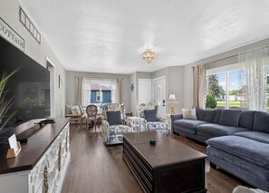 Ein geräumiges Wohnzimmer mit einem dunkelholznen Couchtisch, einem grauen Sofa, gemusterten Sesseln und einem Fernseher auf einem weißen Schrank.