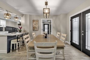 A dining area with a large wooden table, white chairs, pendant lights, and a black-framed door.