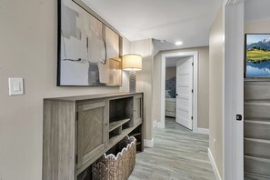 A hallway with a wooden console, a wall-mounted abstract painting, a floor lamp, and a television displaying a mountainous landscape.