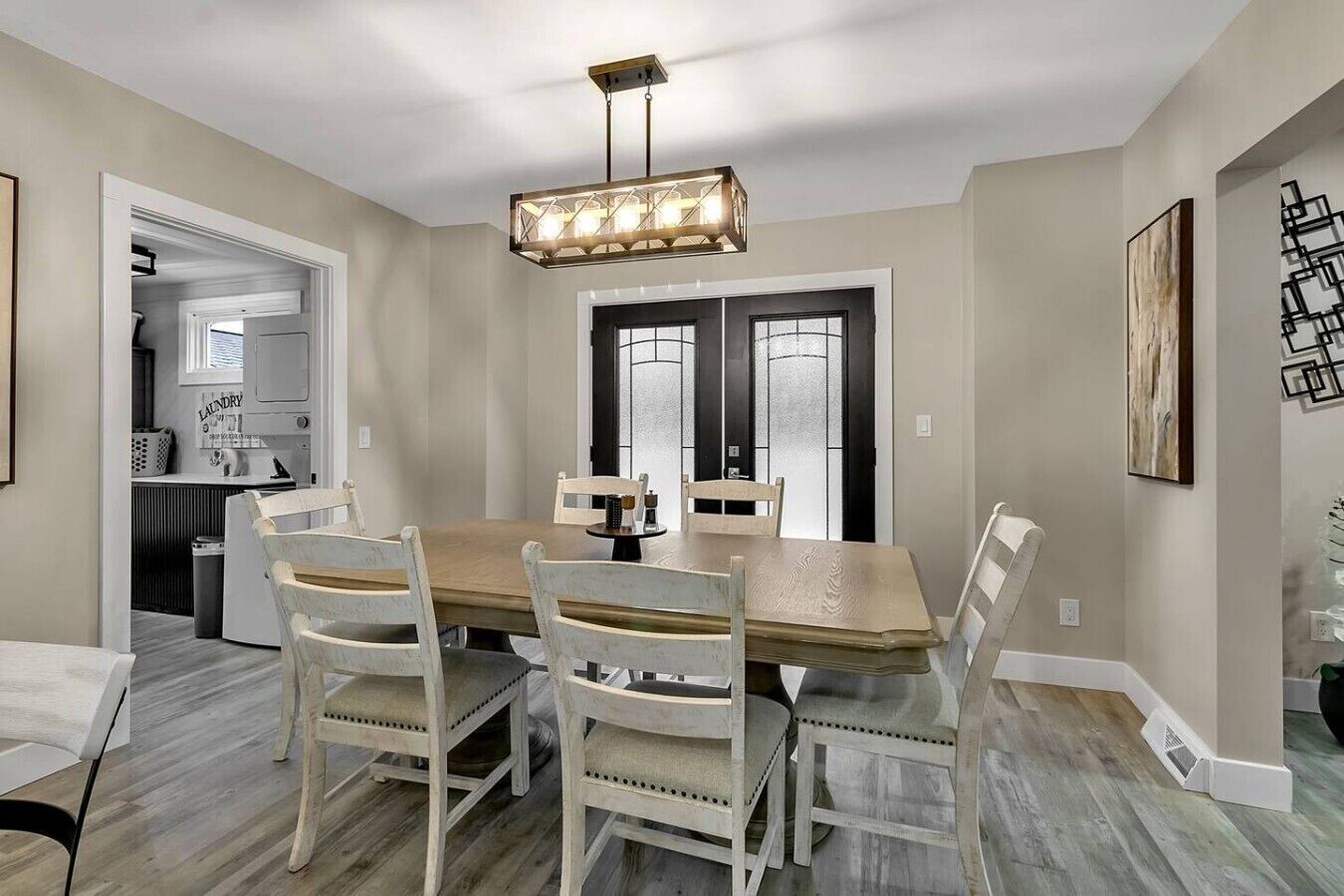 Un espace repas avec une table rectangulaire et six chaises, un luminaire suspendu au-dessus, et une cuisine visible à travers une porte ouverte.
