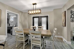 A dining area with a rectangular table and six chairs, a pendant light fixture above, and a kitchen visible through an open door.