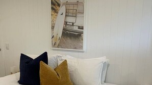 A bedroom with a bed, pillows, a small round table, and a wall-mounted picture of a surfboard. - Killara Apt 1 - Noosa Heads Holiday Apartment (Noosa Heads)