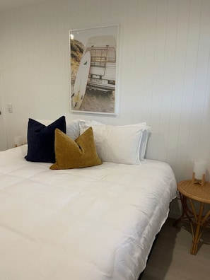 A bedroom with a bed, pillows, a small round table, and a wall-mounted picture of a surfboard. - Killara Apt 1 - Noosa Heads Holiday Apartment (Noosa Heads)