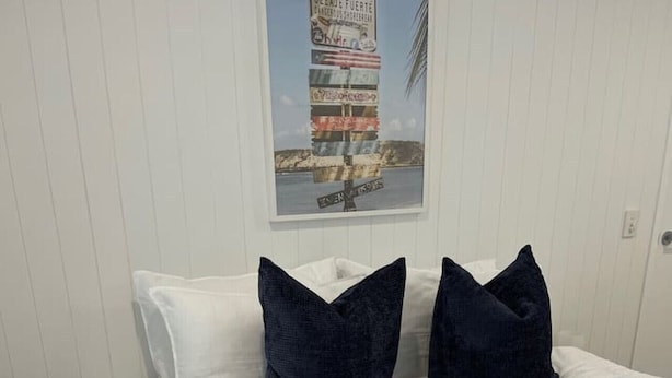 A neatly made bed with white bedding and two dark blue pillows, a small wooden stool, and a framed picture on the wall. - Killara Apt 2 - Noosa Heads Holiday Apartment (Noosa Heads)