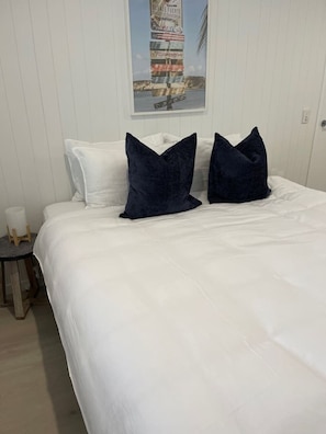 Ein ordentlich bezogenes Bett mit weißer Bettwäsche und zwei dunkelblauen Kissen, ein kleiner Holzschemel und ein gerahmtes Bild an der Wand.