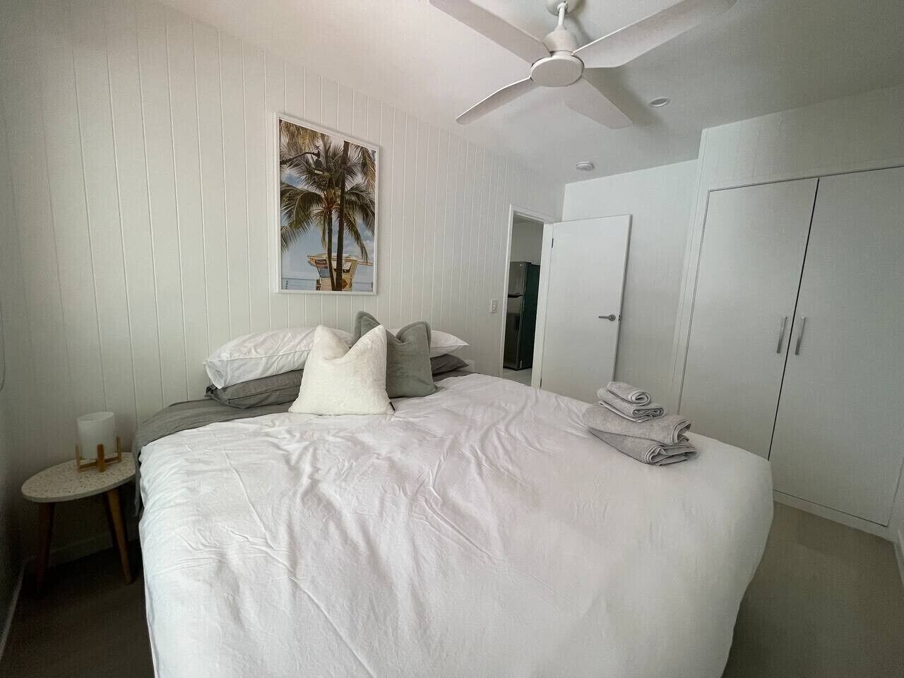 A bedroom with a large bed, a ceiling fan, a painting of palm trees, and a wardrobe.