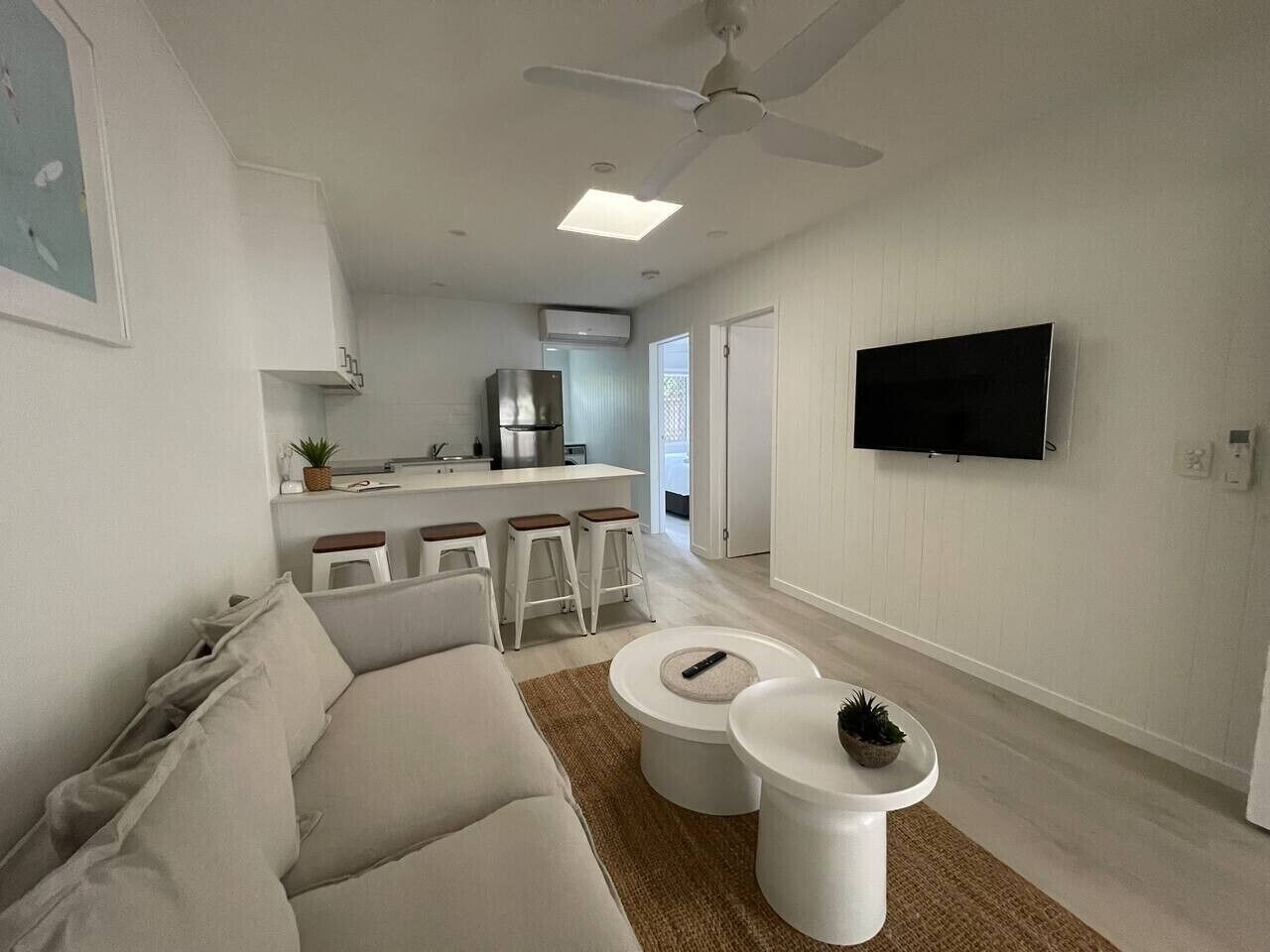 A modern living room with a sofa, two white coffee tables, a television mounted on the wall, and a kitchen area in the background.