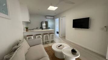 A modern living room with a sofa, two white coffee tables, a television mounted on the wall, and a kitchen area in the background.