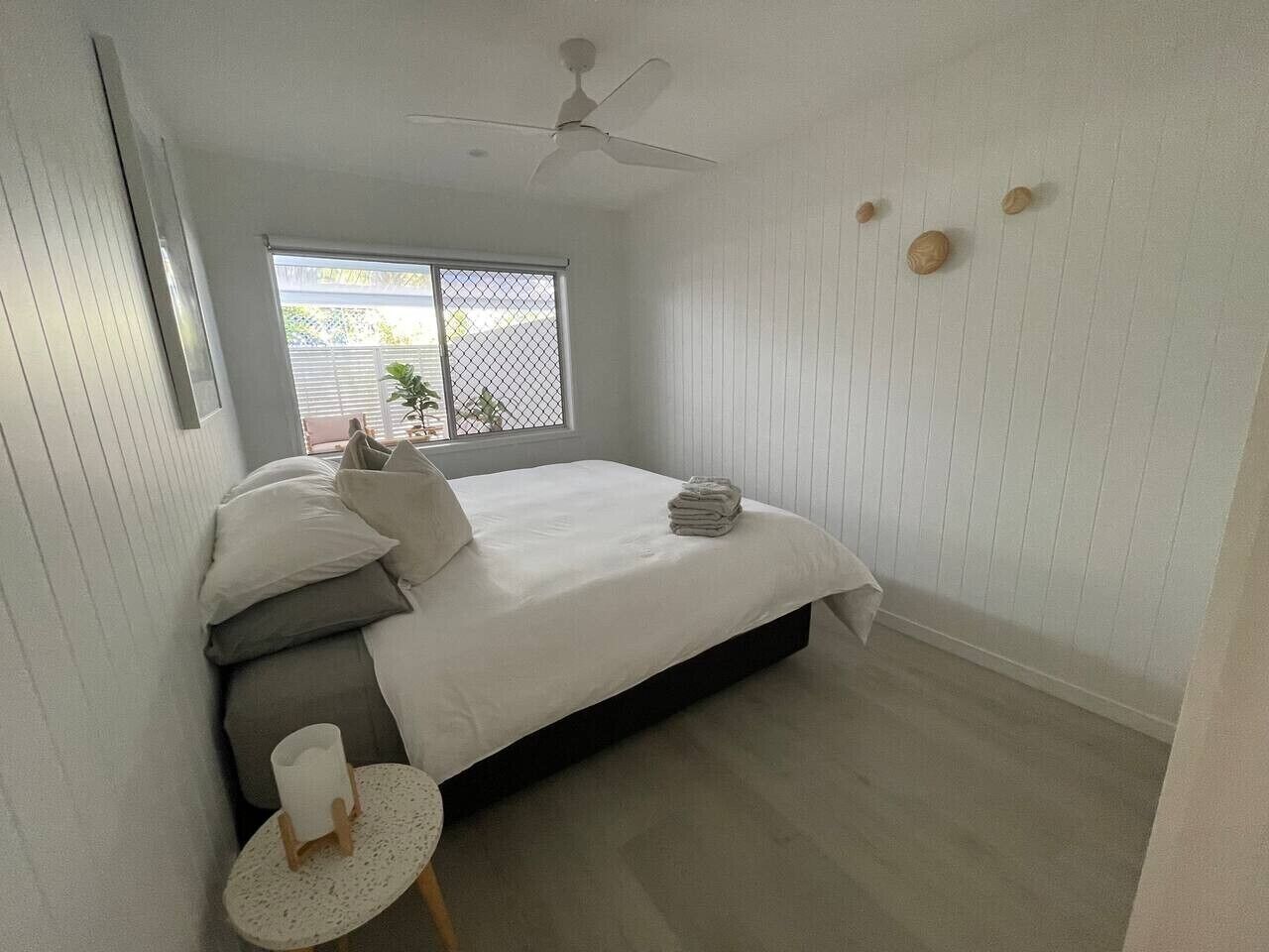 Une chambre à coucher avec un lit, un ventilateur de plafond, une fenêtre avec des stores et une petite table ronde sur laquelle se trouve une tasse.
