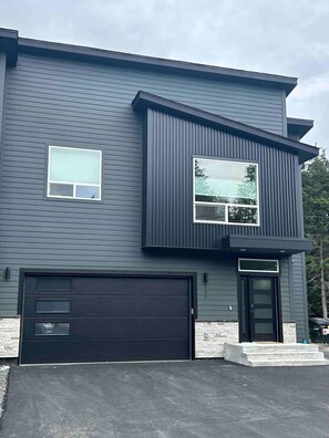 Une maison moderne à deux étages, avec une façade sombre, un garage et une porte d’entrée dotée d’un petit porche.