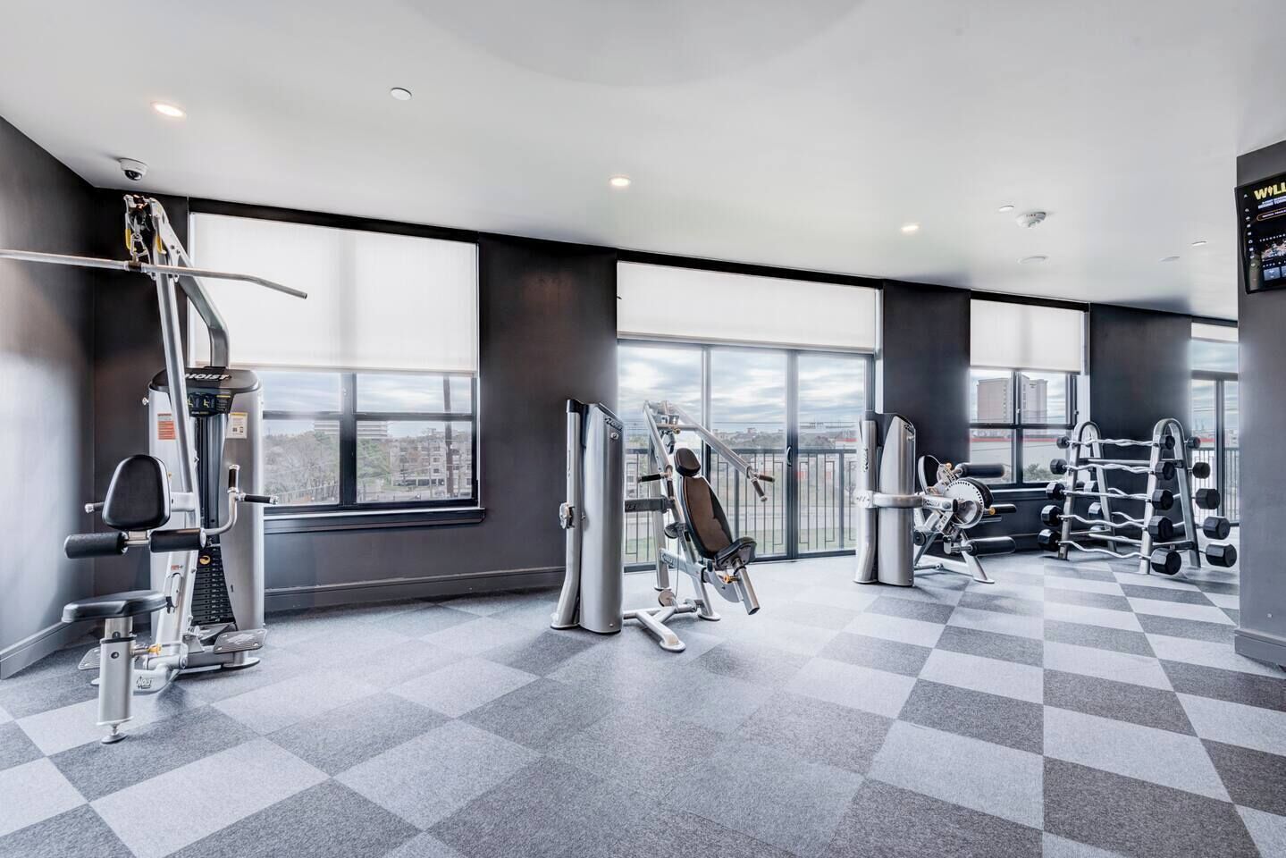 A modern gym with various exercise equipment, large windows, and a checkered floor.