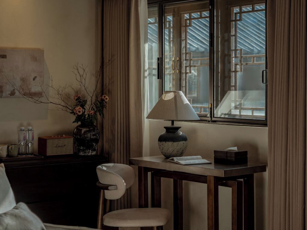 Una habitación con una mesa, una lámpara, una silla, un florero con flores y un ventanal con cortinas.