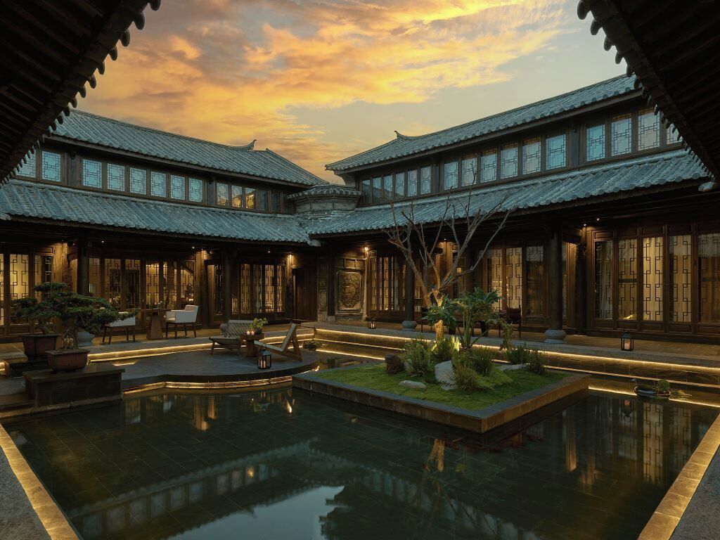Un patio tradicional con un estanque reflectante, edificios de madera con techos de tejas y plantas en macetas.